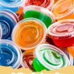 Vibrant rainbow-colored Vodka Jello Shots in small plastic cups, garnished with whipped cream and sprinkles.
