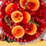 A festive Christmas punch in a large glass bowl with orange slices, lime slices, and cranberries floating on top, with a ladle and glasses nearby.