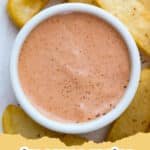 A small glass bowl of creamy orange fry sauce surrounded by golden french fries on a wooden board.