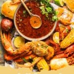 A close-up shot of vibrant orange Seafood Boil Sauce in a glass bowl surrounded by shrimp and corn.